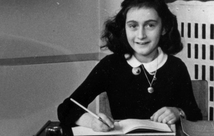 anne frank sitting at a desk and writing in a notebook